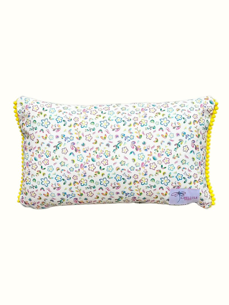 A rectangular lumbar pillow with a dense multicolor floral print on a white background, finished with bright yellow pom-pom trim along the edges.