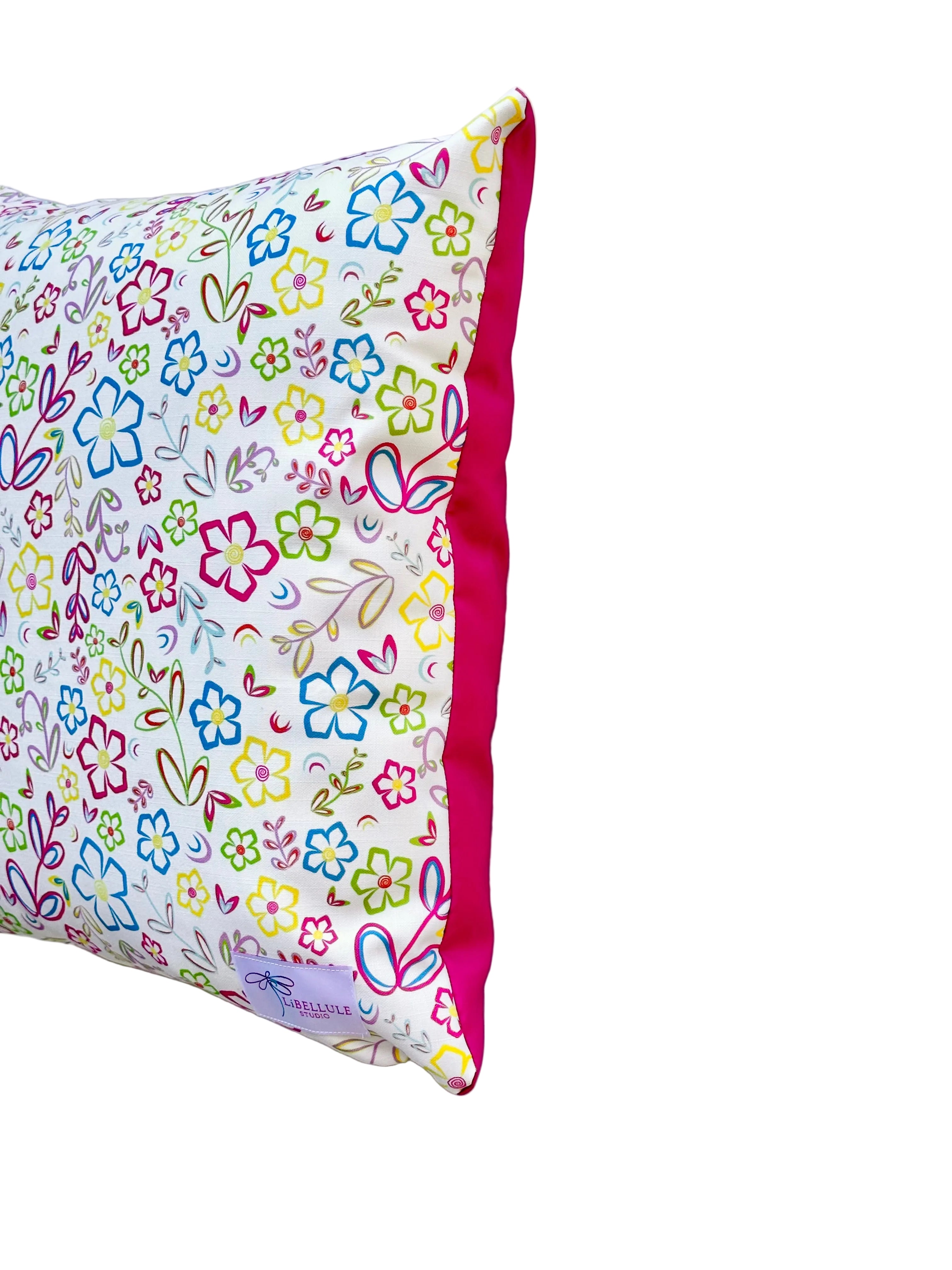 A square outdoor decorative pillow with a colorful floral and butterfly pattern on a white background, finished with a bright pink piped edge, shown against a plain light gray background.