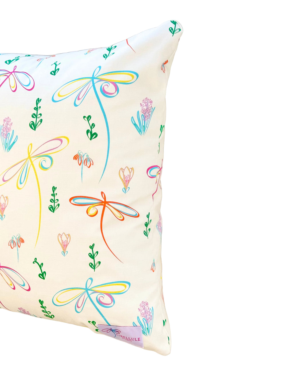 A square throw pillow with a white background and a colorful print of dragonflies and small flowers, shown against a plain beige wall.